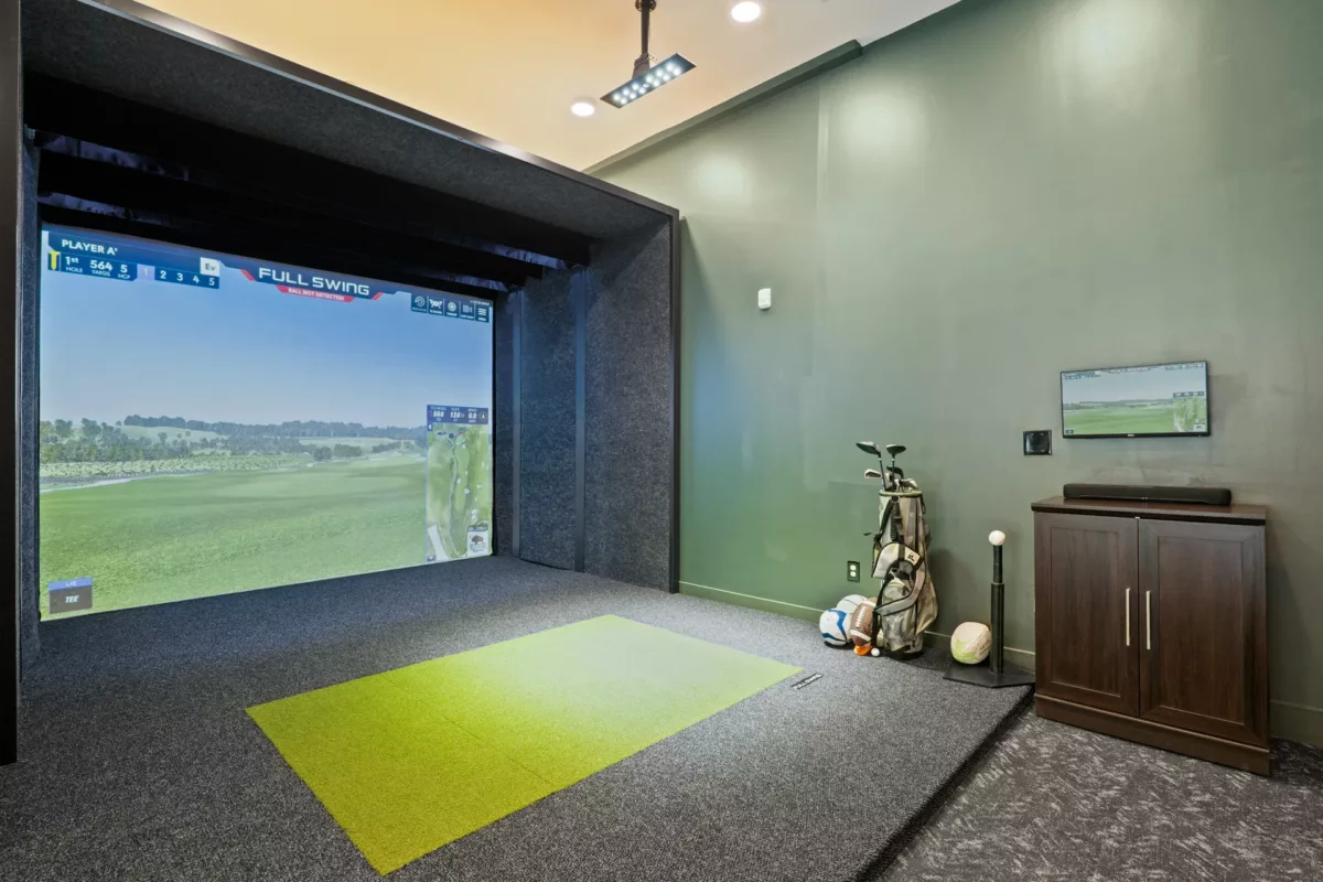Indoor golf simulator room with screen, green turf, golf clubs, and storage cabinet.
