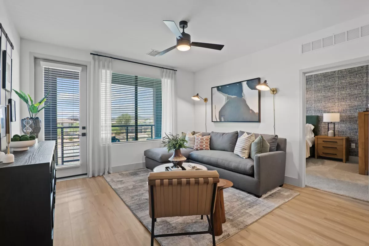 Modern living room with gray sofa, area rug, and view into a bedroom. Ceiling fan present.