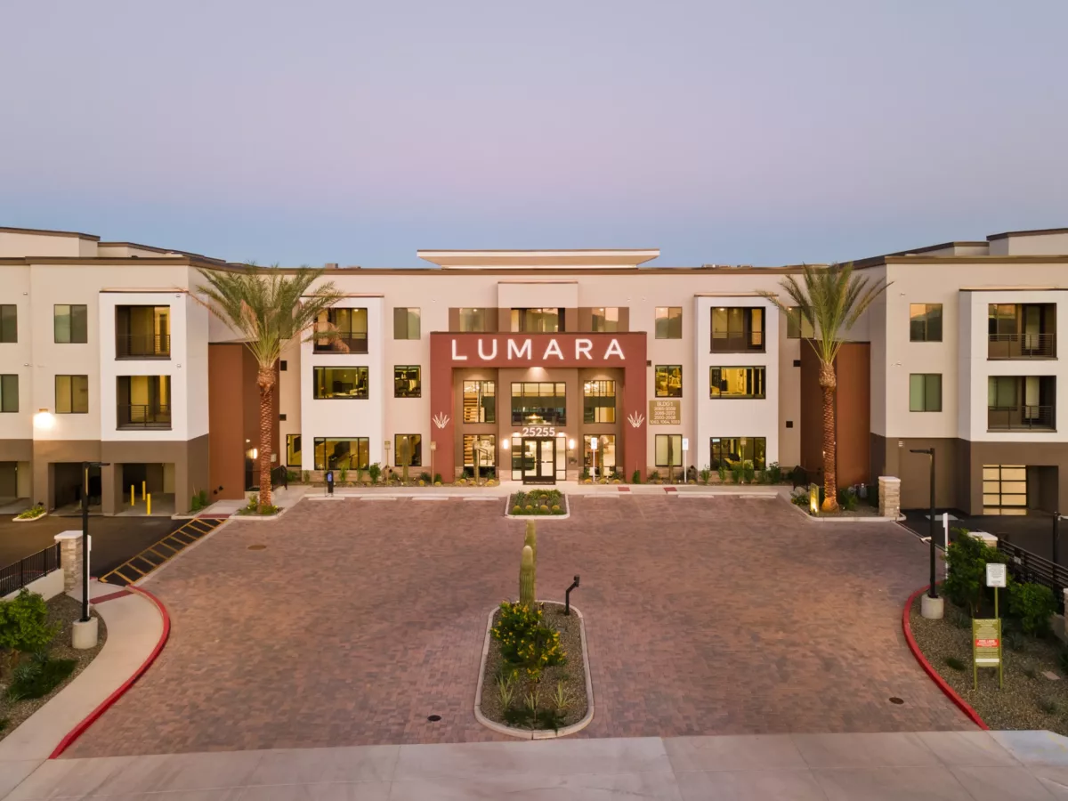 A building exterior with a brick driveway, palm trees, and a central entrance with glass doors under a sign that says "Lumara".