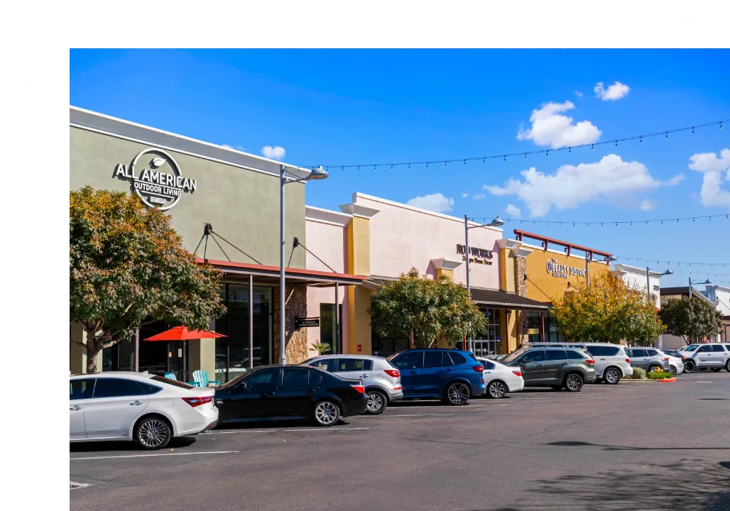 A vibrant shopping center with colorful storefronts, parked cars, and string lights overhead.
